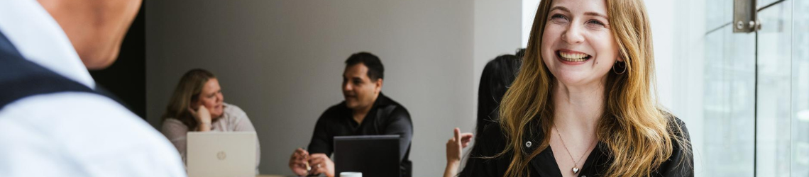 Photo of a woman with long blonde hair smiling and two people in the background using laptops